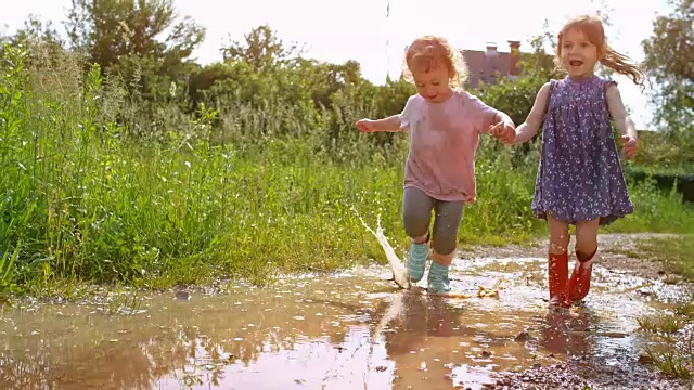 两个小女孩手牵着手，穿着雨靴跑过一个泥泞的水坑视频素材