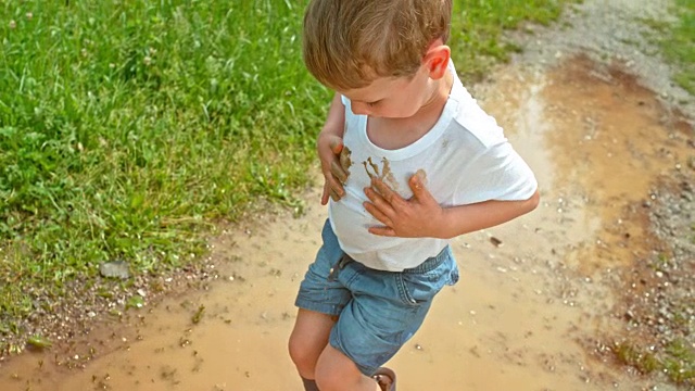 小男孩把他的手放在泥泞的水坑里，用他干净的白色t恤擦拭视频素材