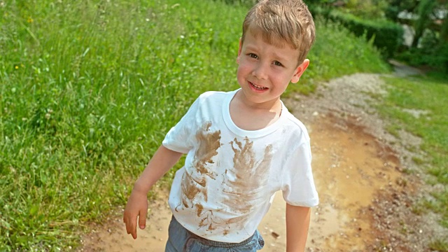 一个穿着脏兮兮的白色t恤的小男孩站在水坑里，对着镜头微笑视频下载