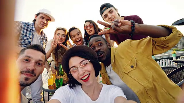 观点拍摄的漂亮的女士戴着时髦的帽子拿着相机和自拍与朋友享受屋顶派对和软饮料，人们看着相机和笑。视频素材