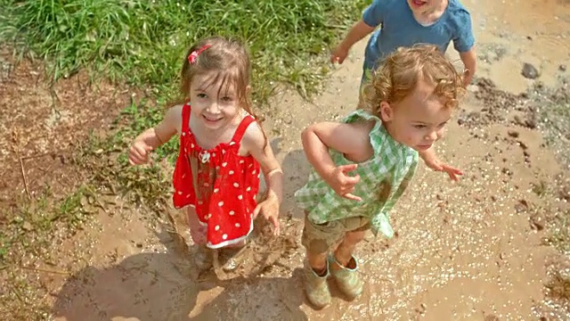 阳光下，三个孩子在泥泞的水坑里跳上跳下视频下载