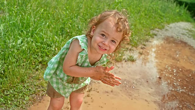 小女孩面带微笑，双手沾满泥巴，站在水坑里，上身脏兮兮的视频下载