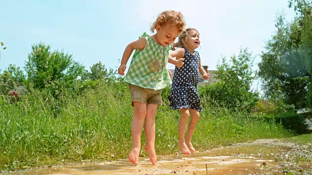 阳光下，两个光着脚的小女孩在泥泞的水坑里跳上跳下视频下载