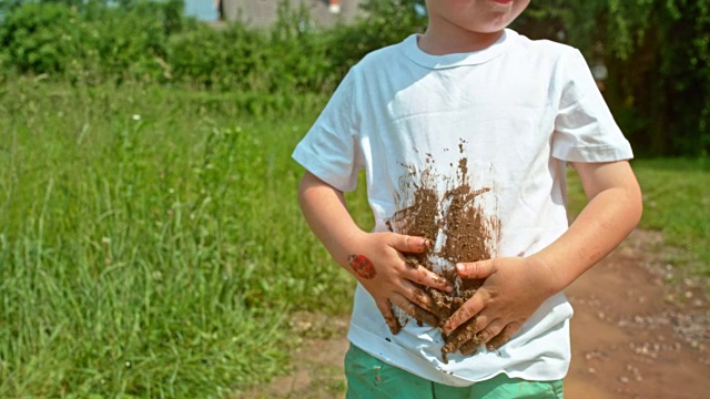 小男孩站在泥泞的水坑里，把泥巴涂在他的白t恤上视频下载