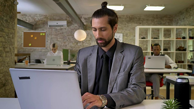 长着胡子的帅气女商人正在办公室的笔记本电脑上打字，看着镜头，微笑着，同事们都在用技术联网，工作理念，交流理念视频素材