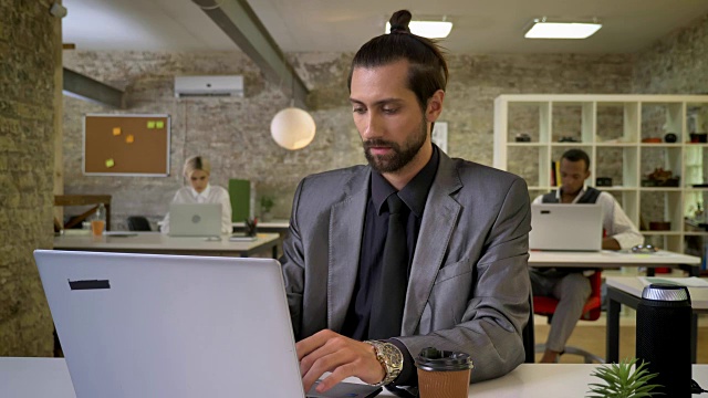 长着胡子的帅气女商人正在办公室的笔记本电脑上打字，喝着咖啡，同事们正在用科技、工作理念、沟通理念进行网络交流视频素材