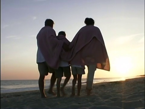 后视图中拍摄的一个家庭走在一起在海滩上手臂在日落。他们身上裹着一条毯子。视频下载