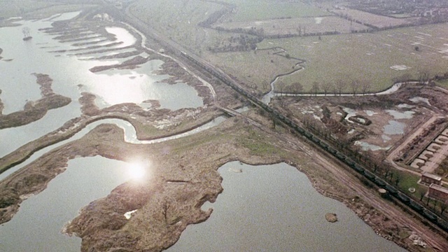 1980年，空中旋转煤运火车沿着水路和开阔的田野轨道运行，在朦胧的天空下，陆地分叉成水道，叙述者在讨论火车和时刻表/英国视频素材