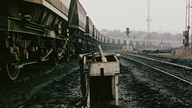 1980年蒙太奇煤炭火车缓慢地在轨道上移动，在一个悬垂的站台下，旁边是信号，背景是金属塔，旁白正在讨论煤炭运输/英国视频素材